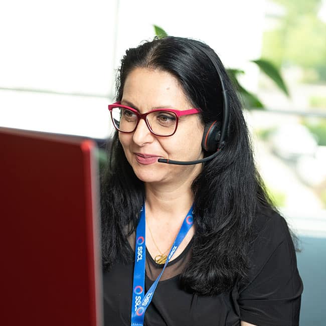 Lady in office, wearing glasses, on headset,looking at PC screen.