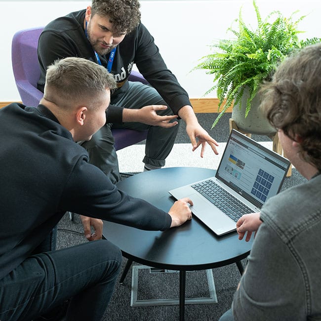 Three-men-sat-round-a-pc-on-small-round-table,-looking-at-screen